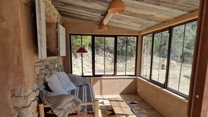A cozy room with a stone wall, a wooden bench with cushions, a patterned rug, and large windows offering a view of the outdoors.