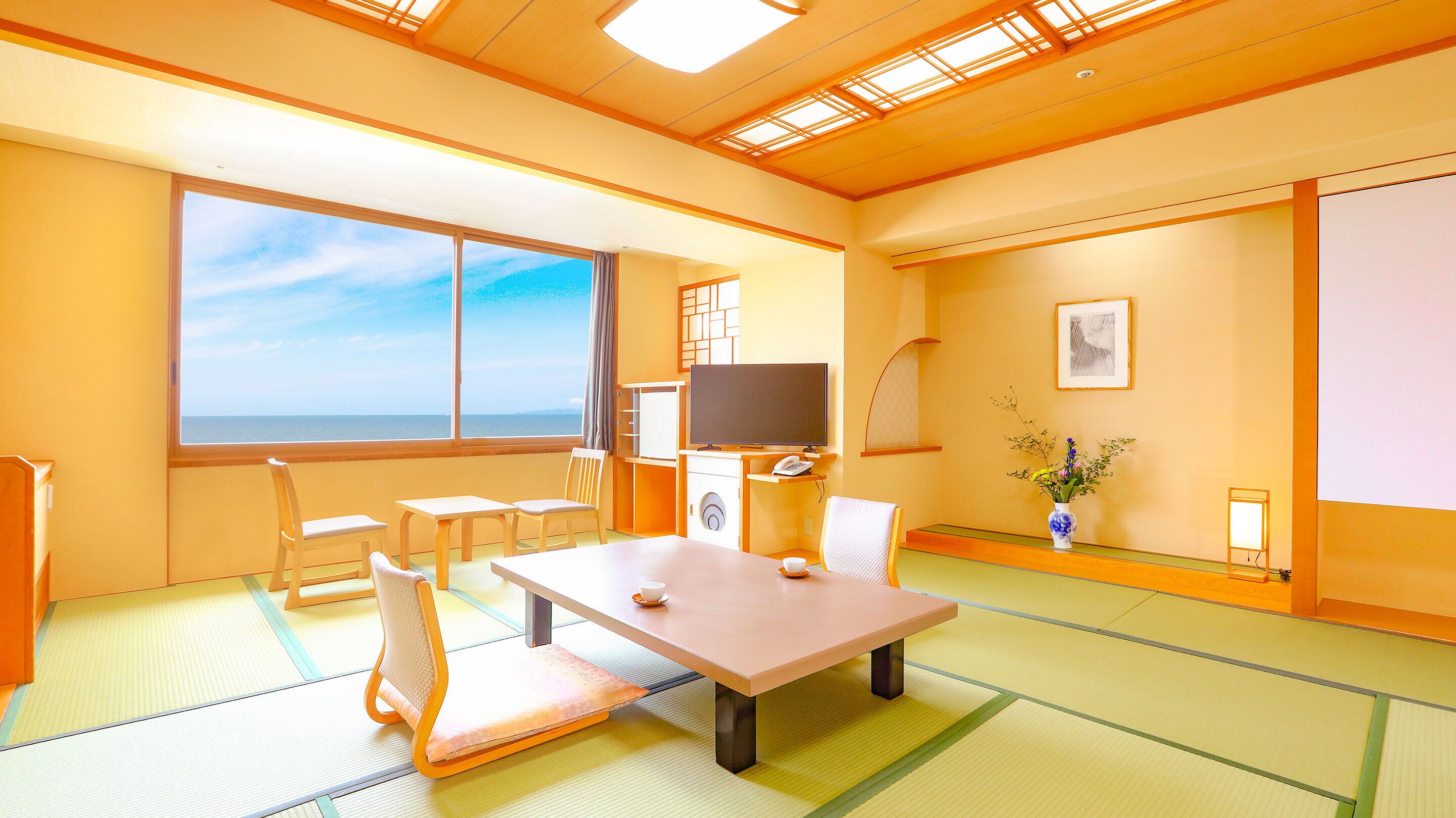 A room with tatami mats, a table, chairs, a television, a telephone, a vase with flowers, and a view of the sea.