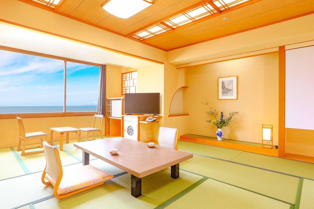 Una habitación con esteras de tatami, una mesa, sillas, un televisor, un teléfono, un jarrón con flores y vista al mar.