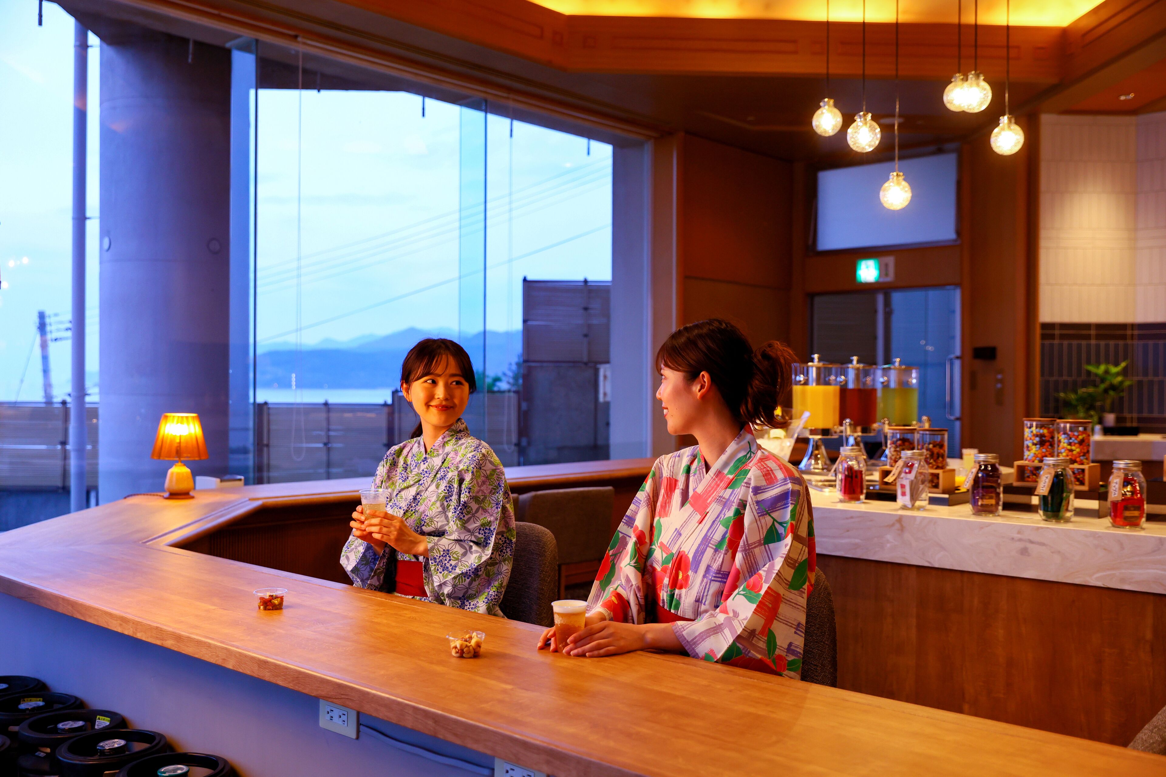 A bar area with two people seated at a counter, a lamp, and various bottles and jars on the counter.