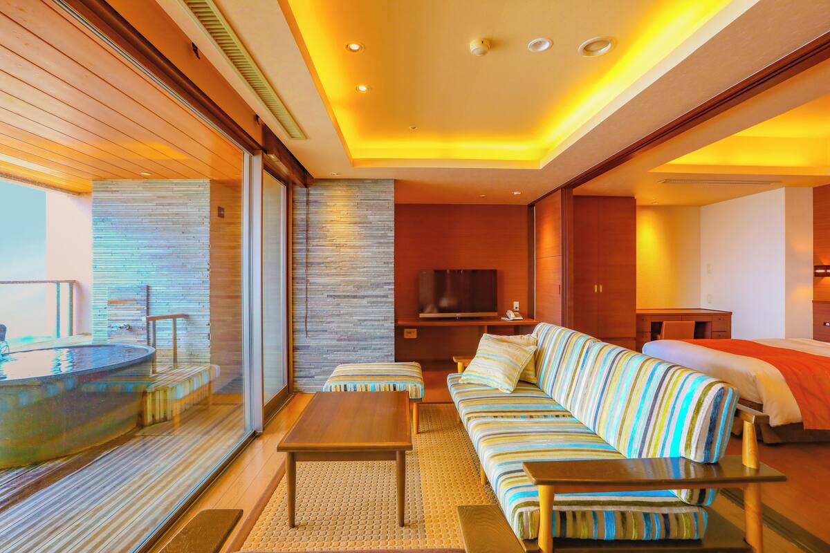 Habitación de hotel con una cama grande, un sofá, un televisor y una mesita. Se ve una ventana con vistas al exterior.