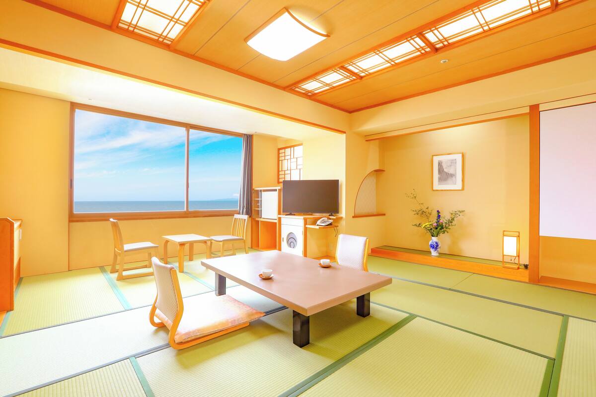 Una habitación japonesa tradicional con suelo de tatami, una mesa baja, sillas, un televisor y un ventanal con vistas al cielo.