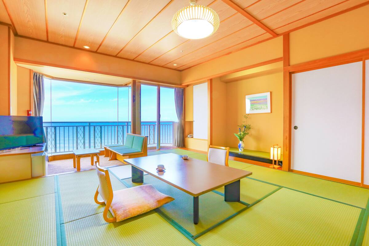 Una habitación japonesa tradicional con suelo de tatami, una mesa baja, sillas, televisor y un balcón con vistas.