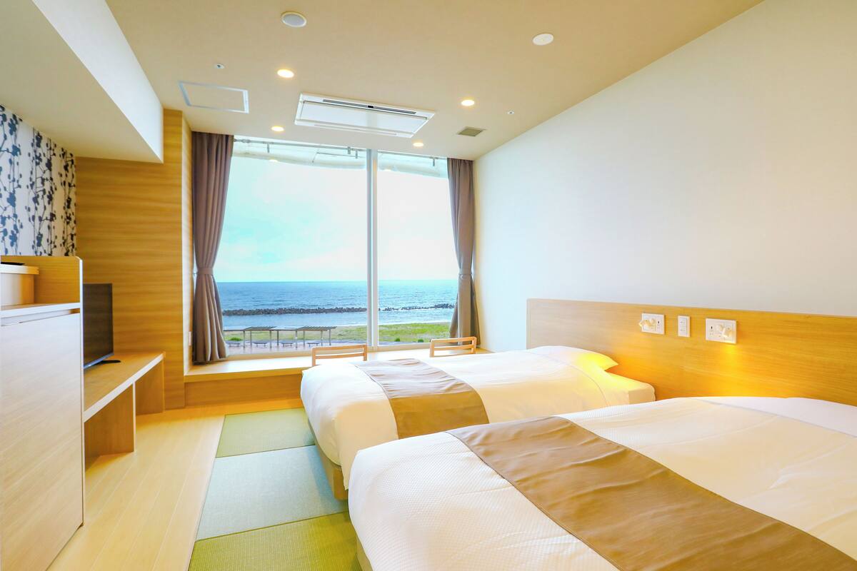 Habitación de hotel con dos camas, un televisor, un ventanal con vista al mar y un ventilador de techo.