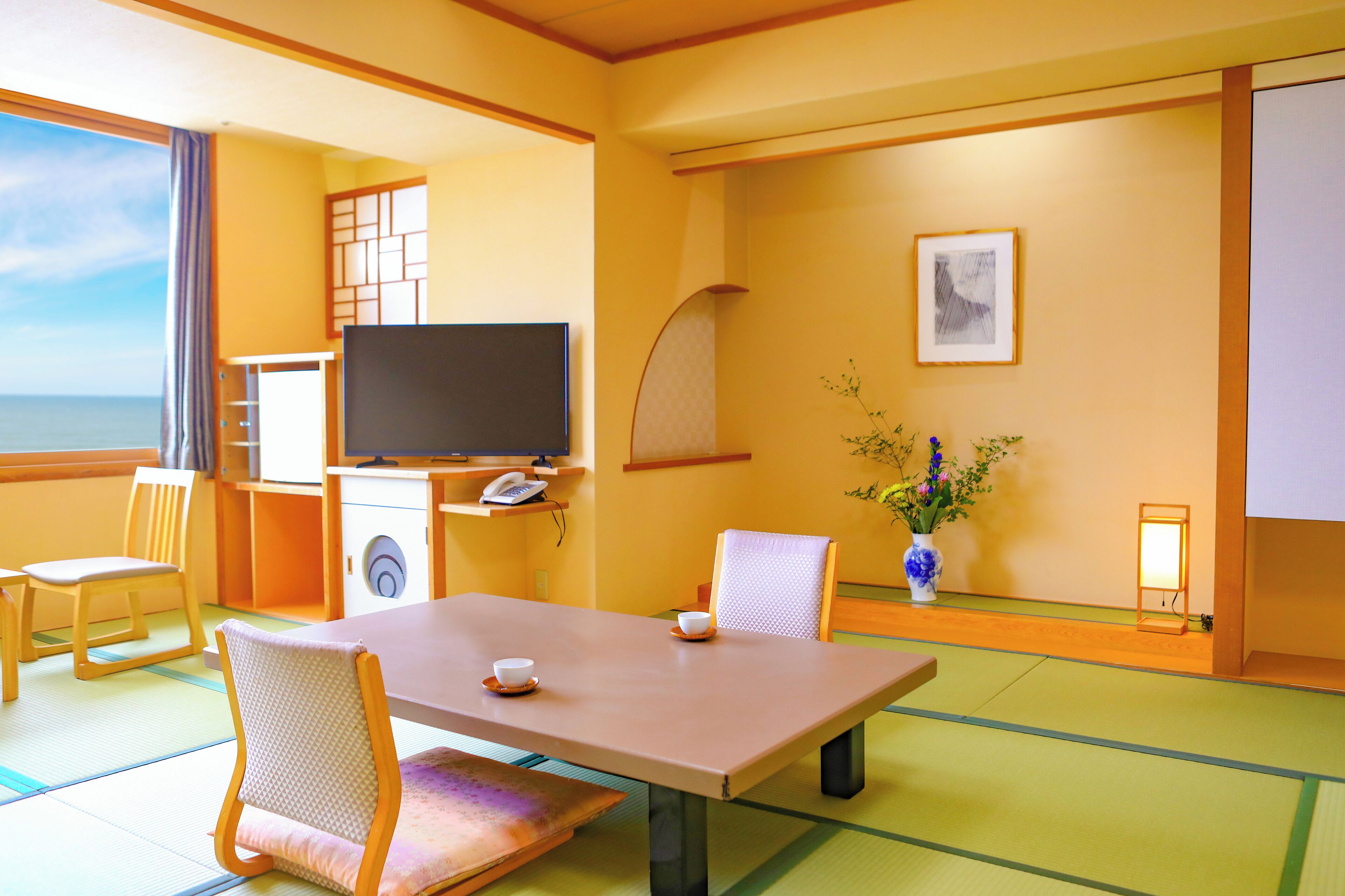 Una habitación de estilo japonés tradicional con suelo de tatami, una mesa baja, sillas, un televisor y un jarrón con flores.