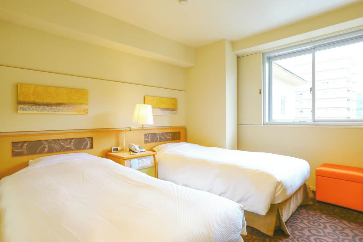 Habitación de hotel con dos camas, una mesita de noche con lámpara, teléfono y ventana con vista a edificios.