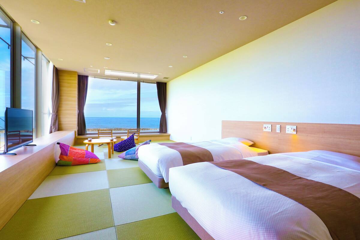 Habitación de hotel con dos camas, televisión, ventana con vista al mar y una pequeña mesa con una silla.