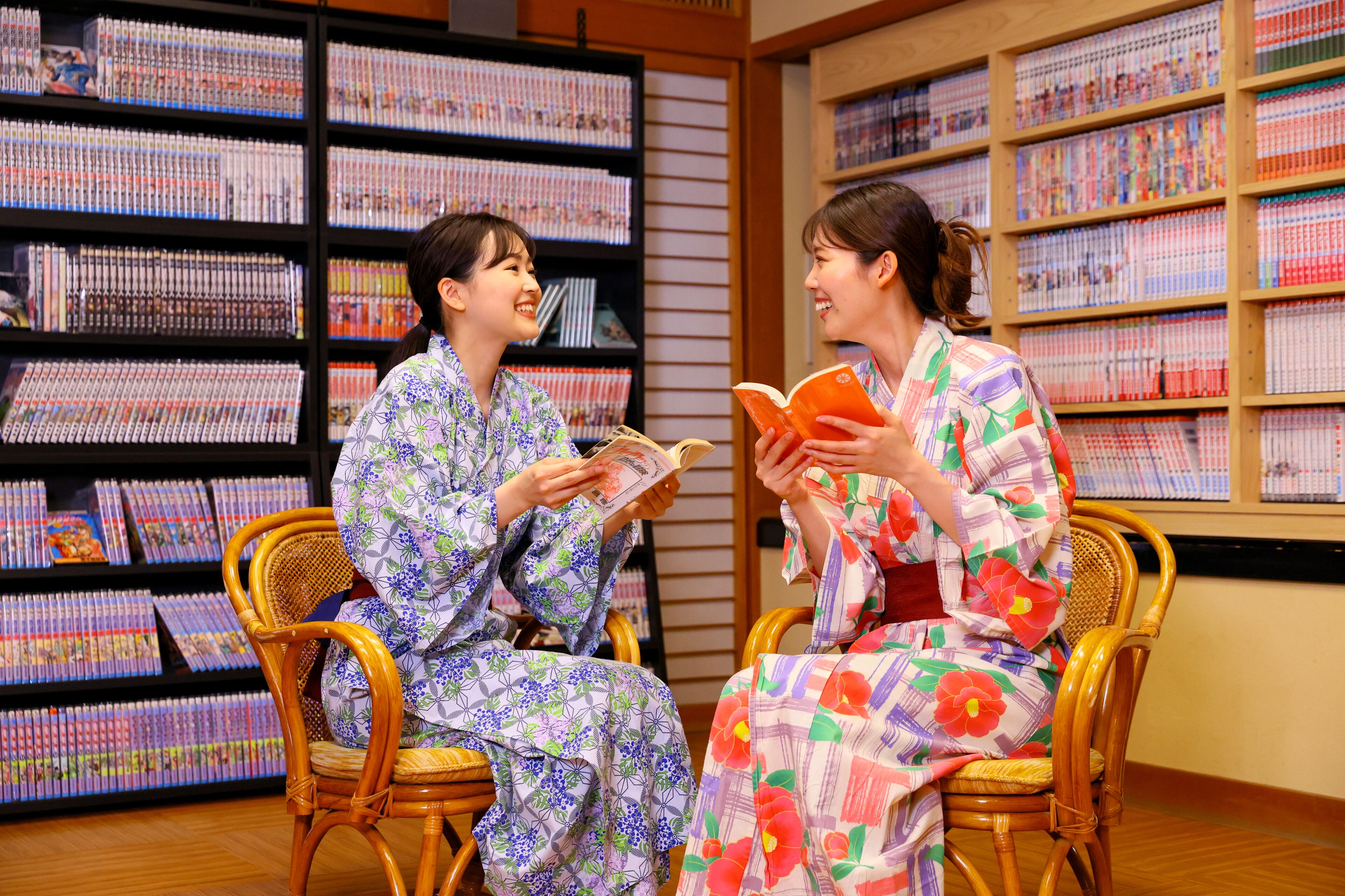 Two individuals seated in wicker chairs, reading books in a room with wooden flooring and shelves filled with books.