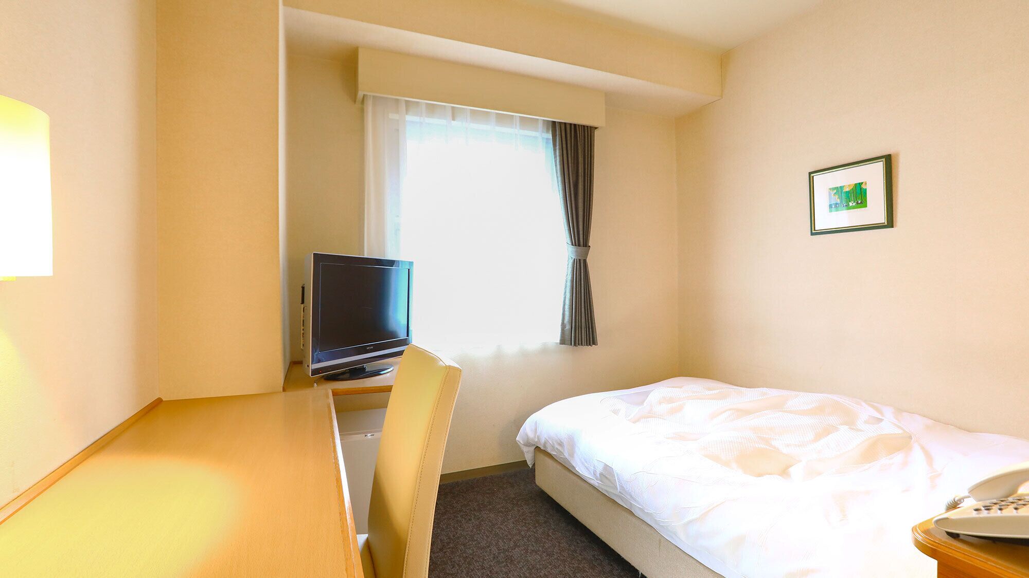 A hotel room with a bed, a desk with a laptop, a window with curtains, and a framed picture on the wall.