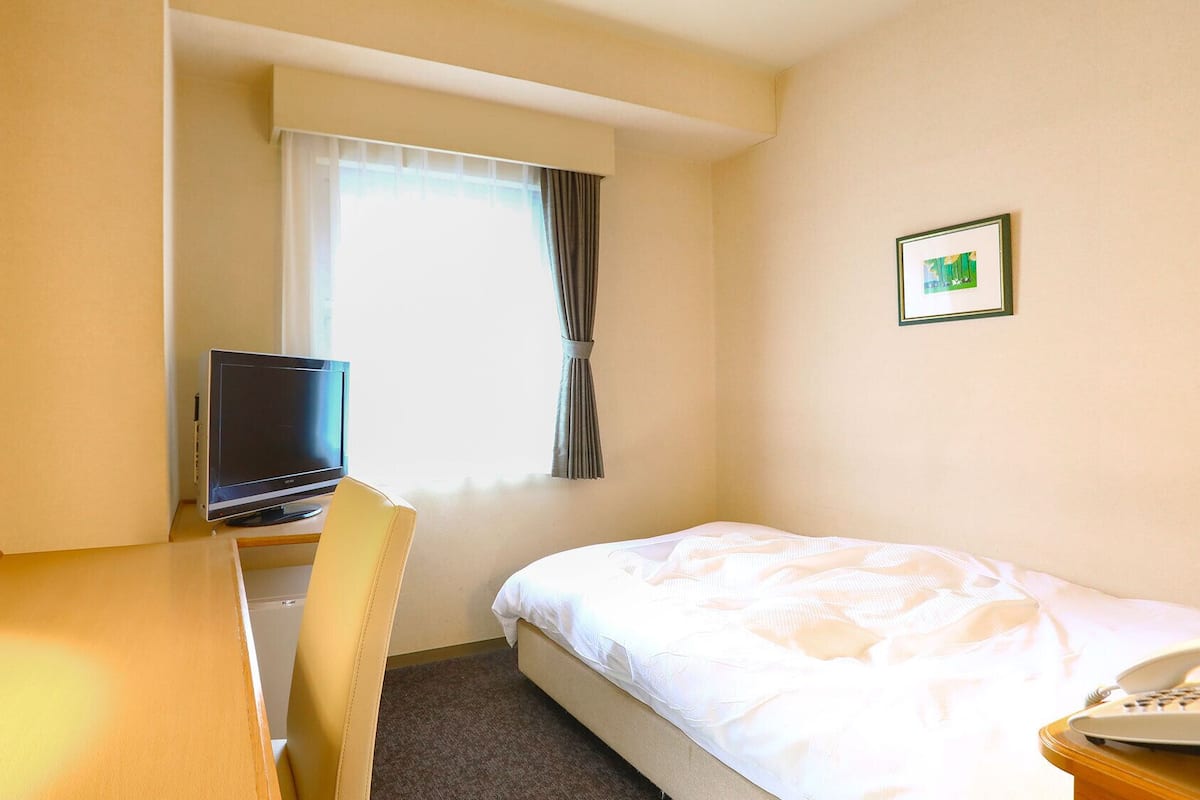 Una habitación de hotel con cama, un escritorio con computadora portátil, una ventana con cortinas y un cuadro en la pared.