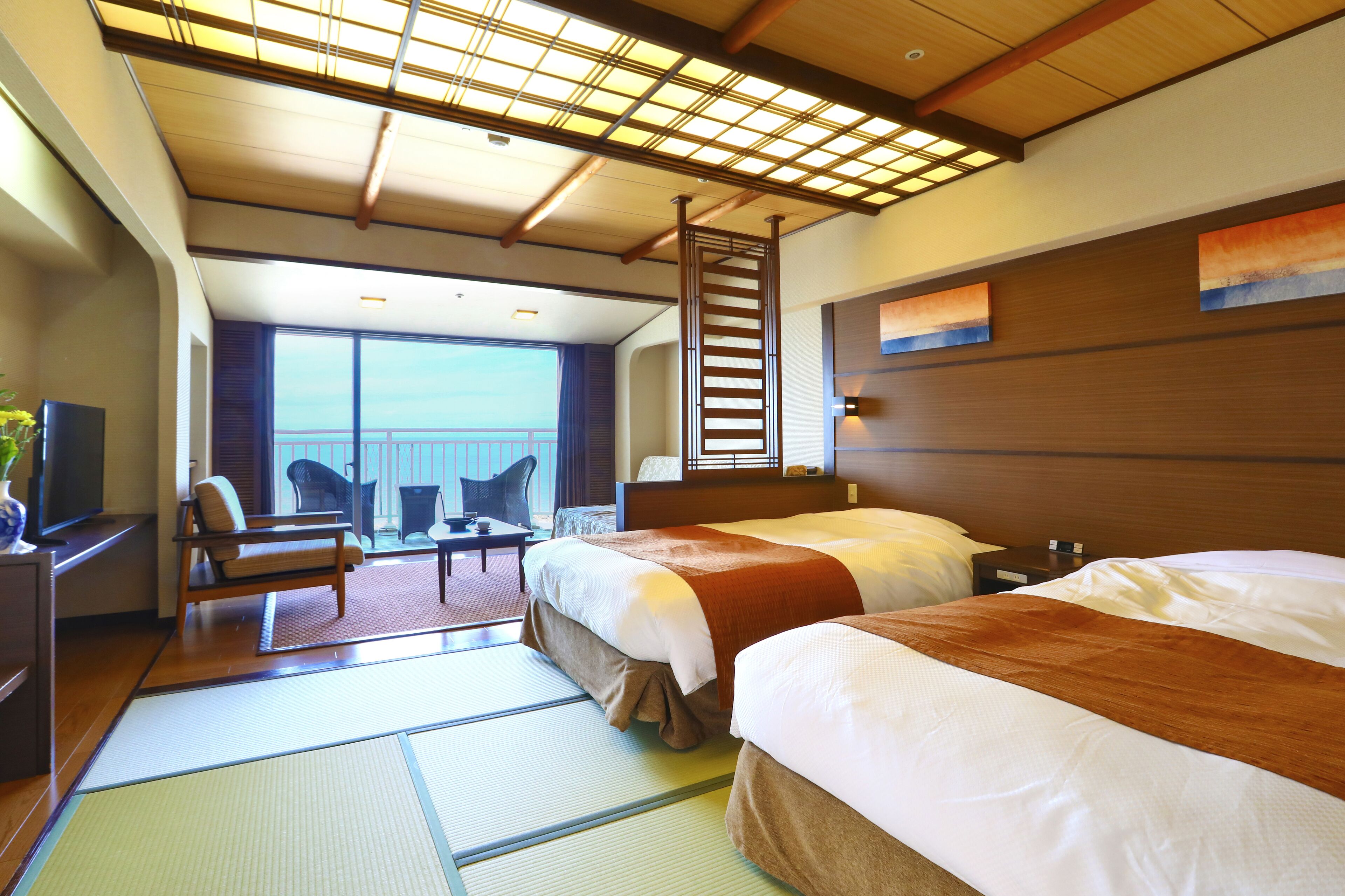 A hotel room with two beds, a sitting area with a chair and table, a TV, and a view of the sea.