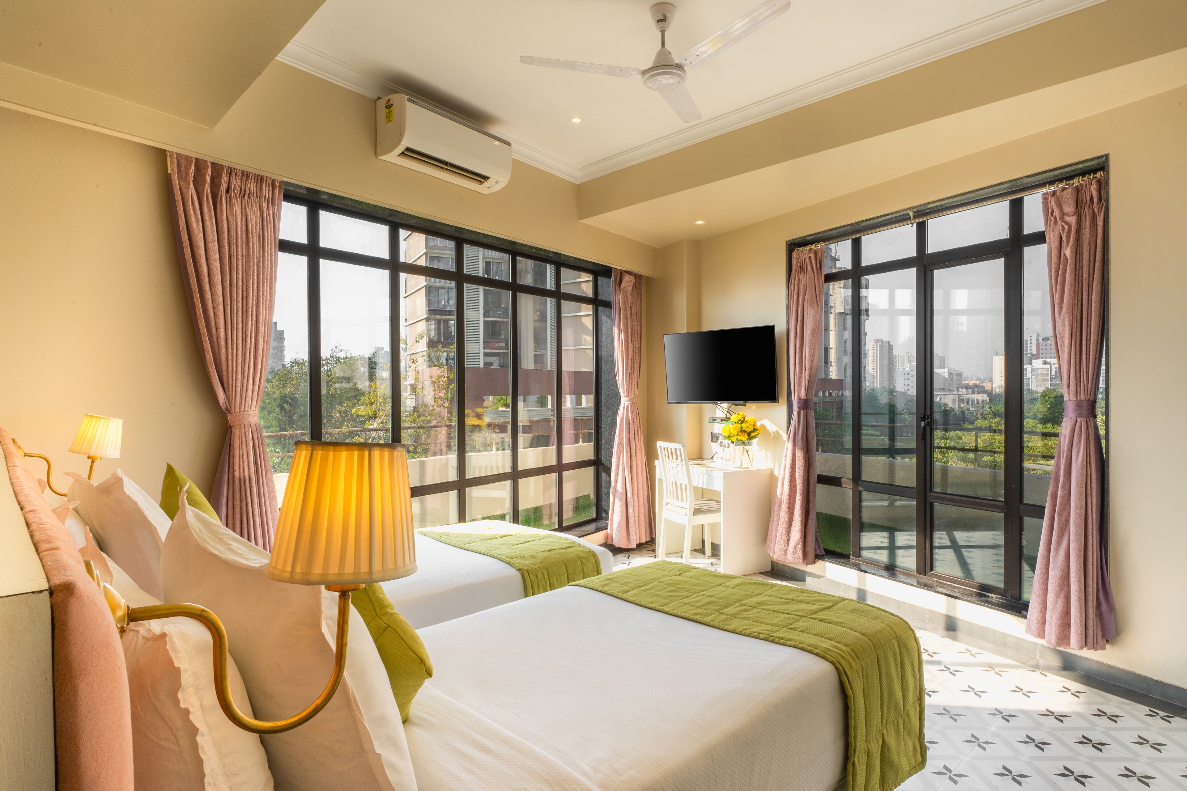 Una habitación de hotel con dos camas, televisión, escritorio con silla y ventanas amplias con cortinas.