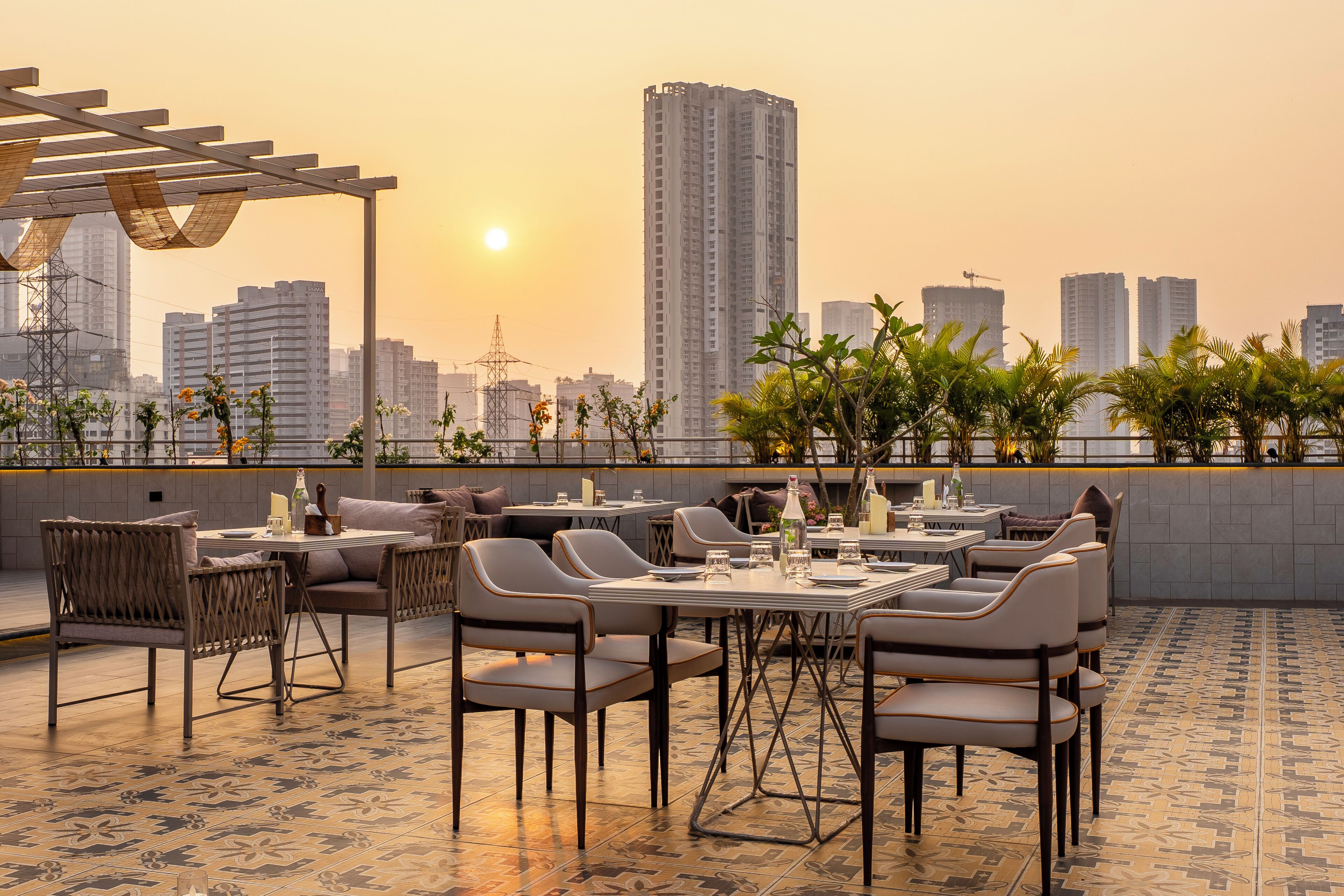 An outdoor dining area with tables and chairs, a pergola, and a city skyline in the background.