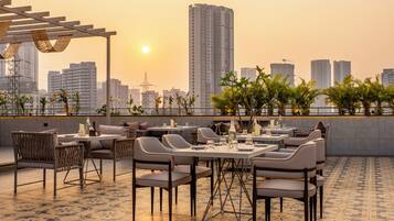 An outdoor dining area with tables and chairs, a pergola, and a city skyline in the background.