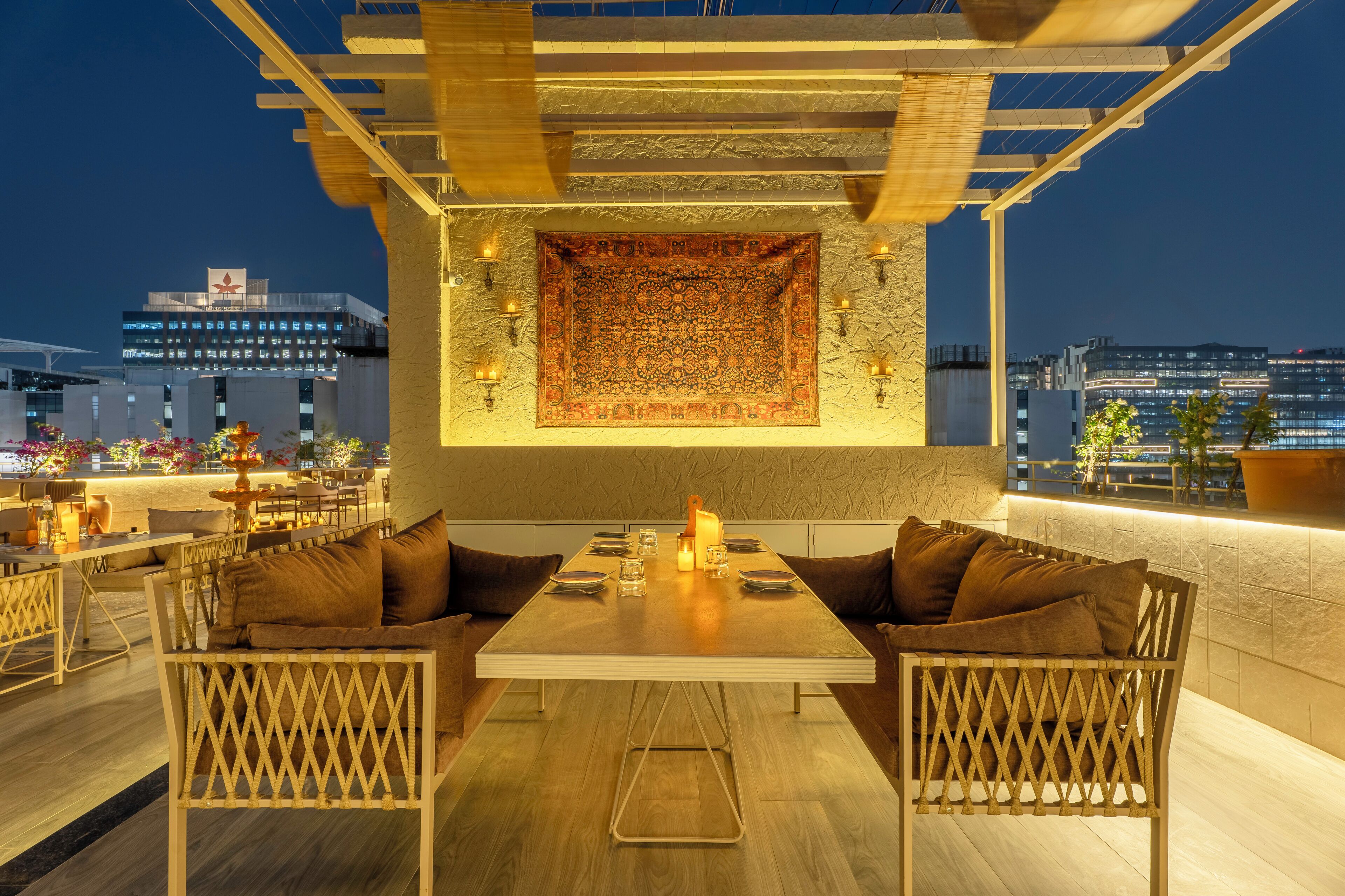 An outdoor seating area with a table and chairs, a patterned wall hanging, and a cityscape view in the background.
