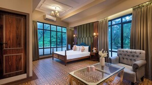 Une chambre d’hôtel dotée d’un grand lit, d’un coin salon avec un fauteuil, d’une table basse en verre et offrant une vue sur la verdure grâce à de grandes fenêtres.