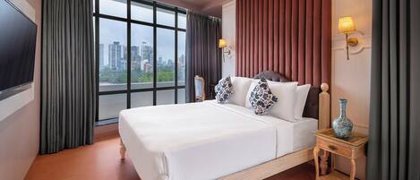 Habitación de hotel con una cama grande, televisión, ventana con cortinas, mesita de noche con un florero y vista a la ciudad.