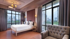 Une chambre d’hôtel avec un grand lit, un canapé, une table basse avec des tasses, et une vue sur la ville grâce à de grandes fenêtres.