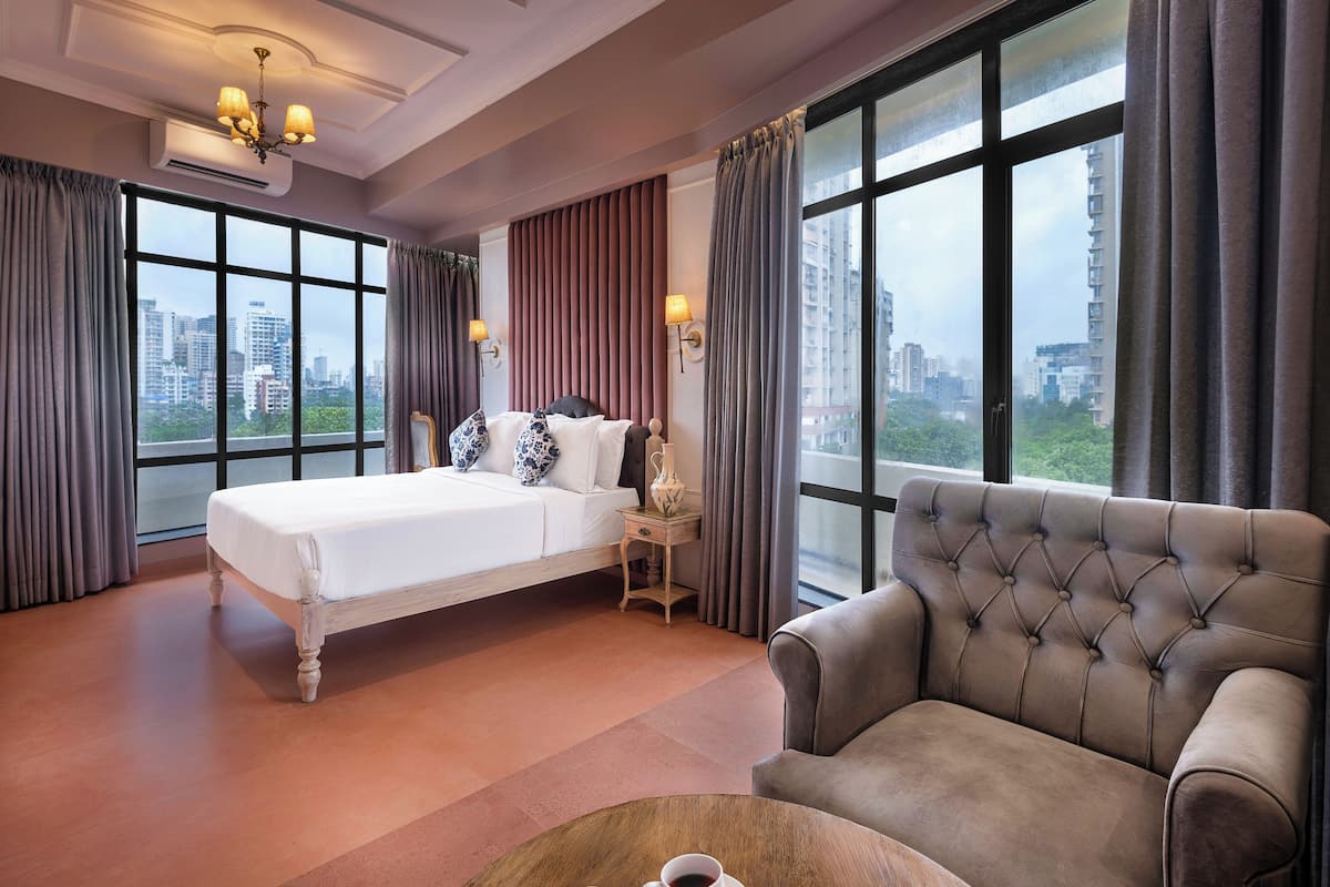 Quarto de hotel com uma cama grande, um sofá, uma mesa de centro com chávenas e vista para a cidade através de amplas janelas.