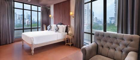 A hotel room with a large bed, a sofa, a coffee table with cups, and a view of the city through large windows.