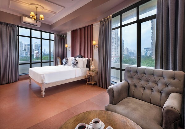 A hotel room with a large bed, a sofa, a coffee table with cups, and a view of the city through large windows. - Arnna Hotel - Goregaon (Mumbai)