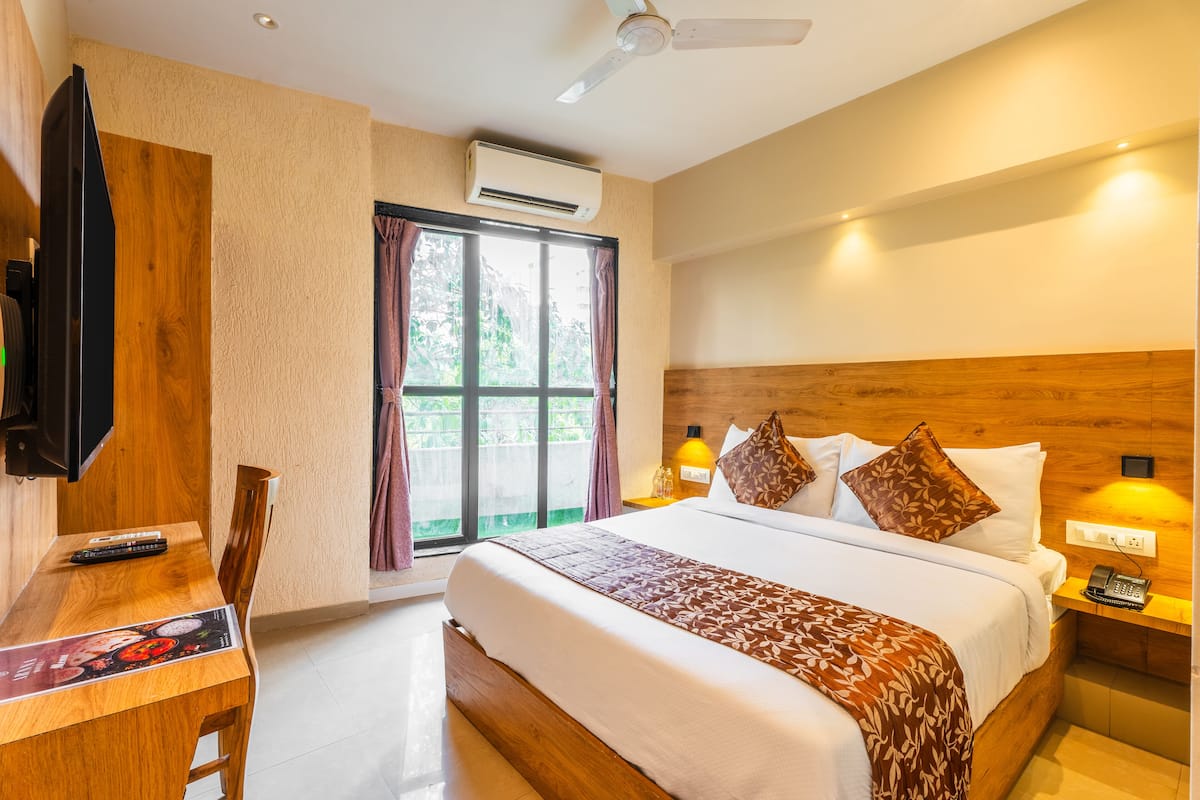 Quarto de hotel com cama, escrivaninha com televisão, janela com cortinas e ventilador de teto.