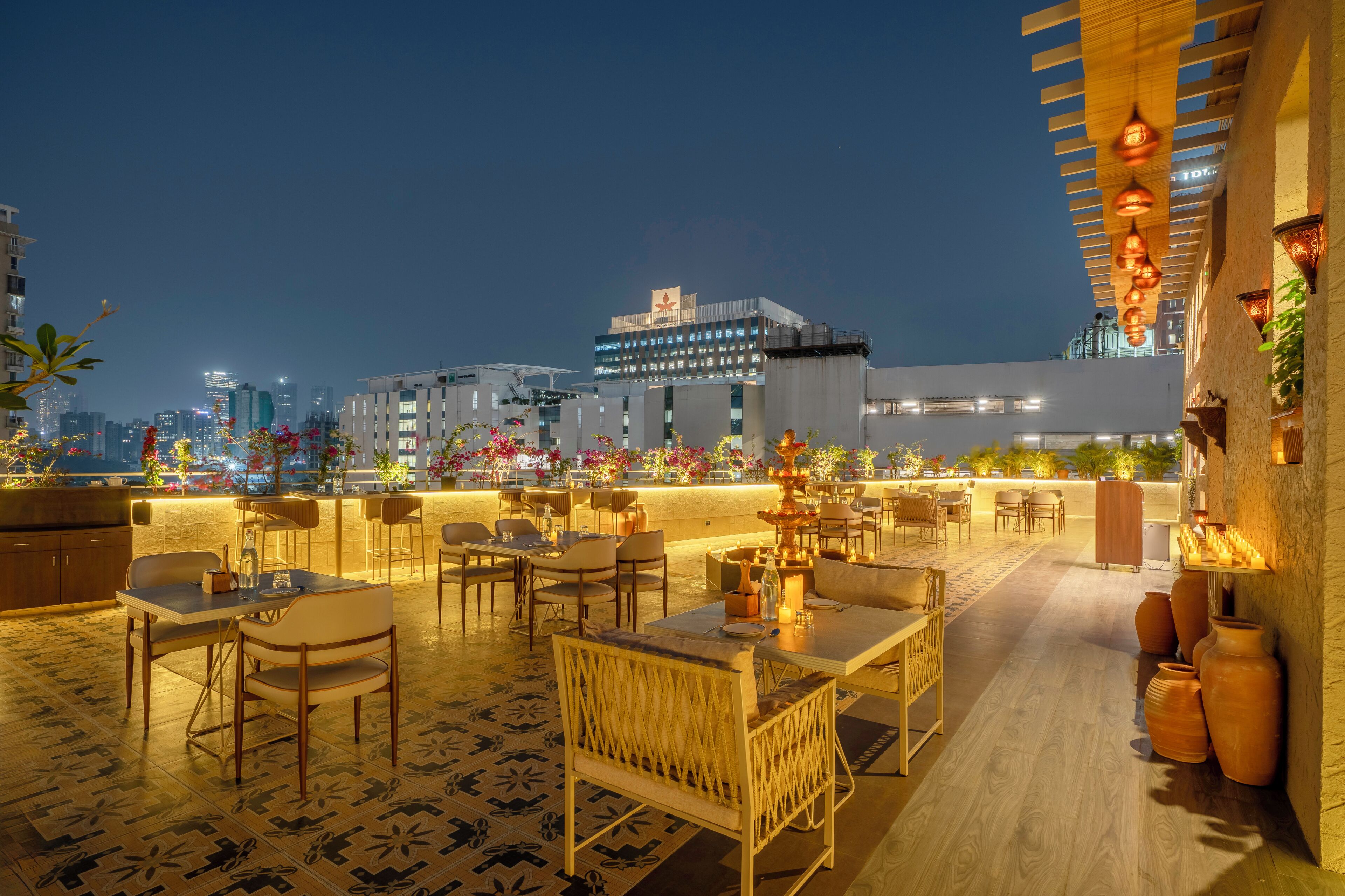 An outdoor dining area with tables and chairs, a decorative fountain, and a view of the city skyline at night.