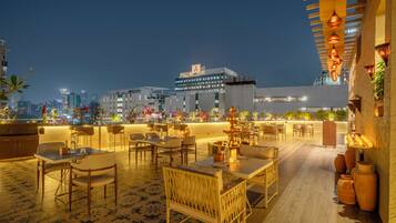 An outdoor dining area with tables and chairs, a decorative fountain, and a view of the city skyline at night.