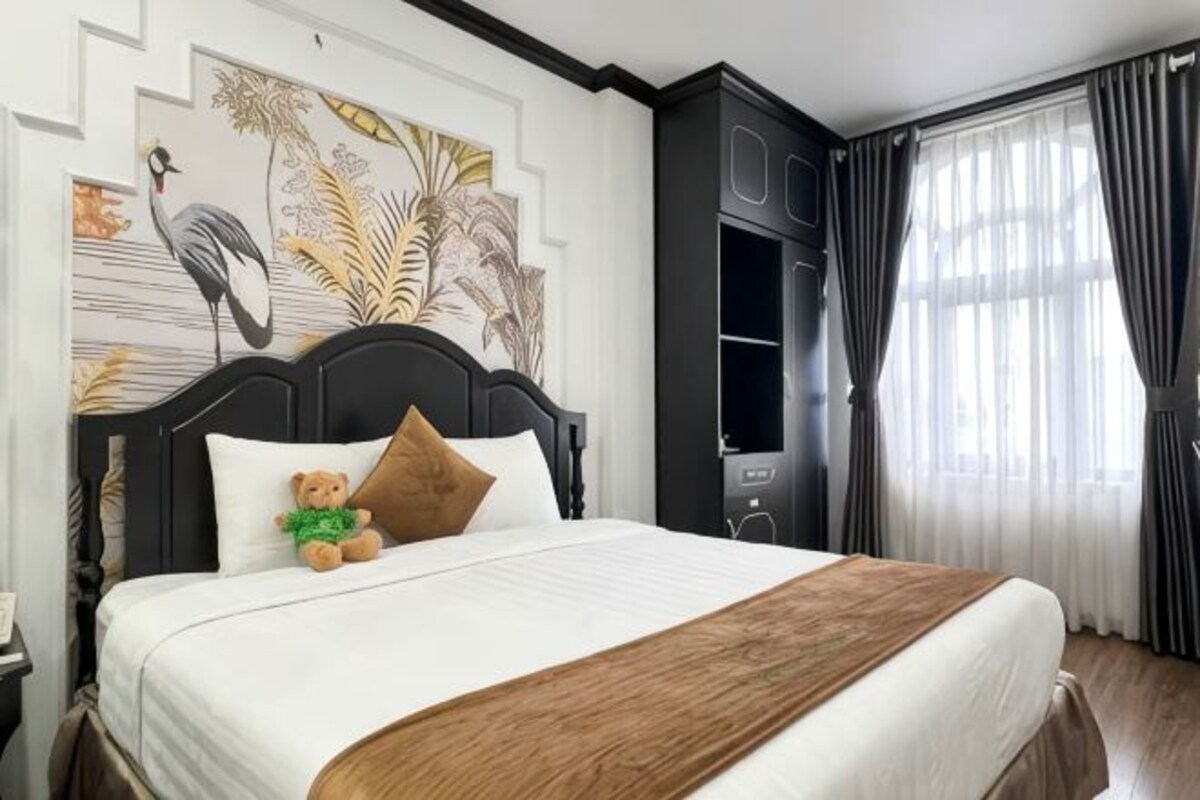 A hotel room with a bed, a nightstand, a wall with a bird and plant mural, a window with curtains, and a wardrobe.