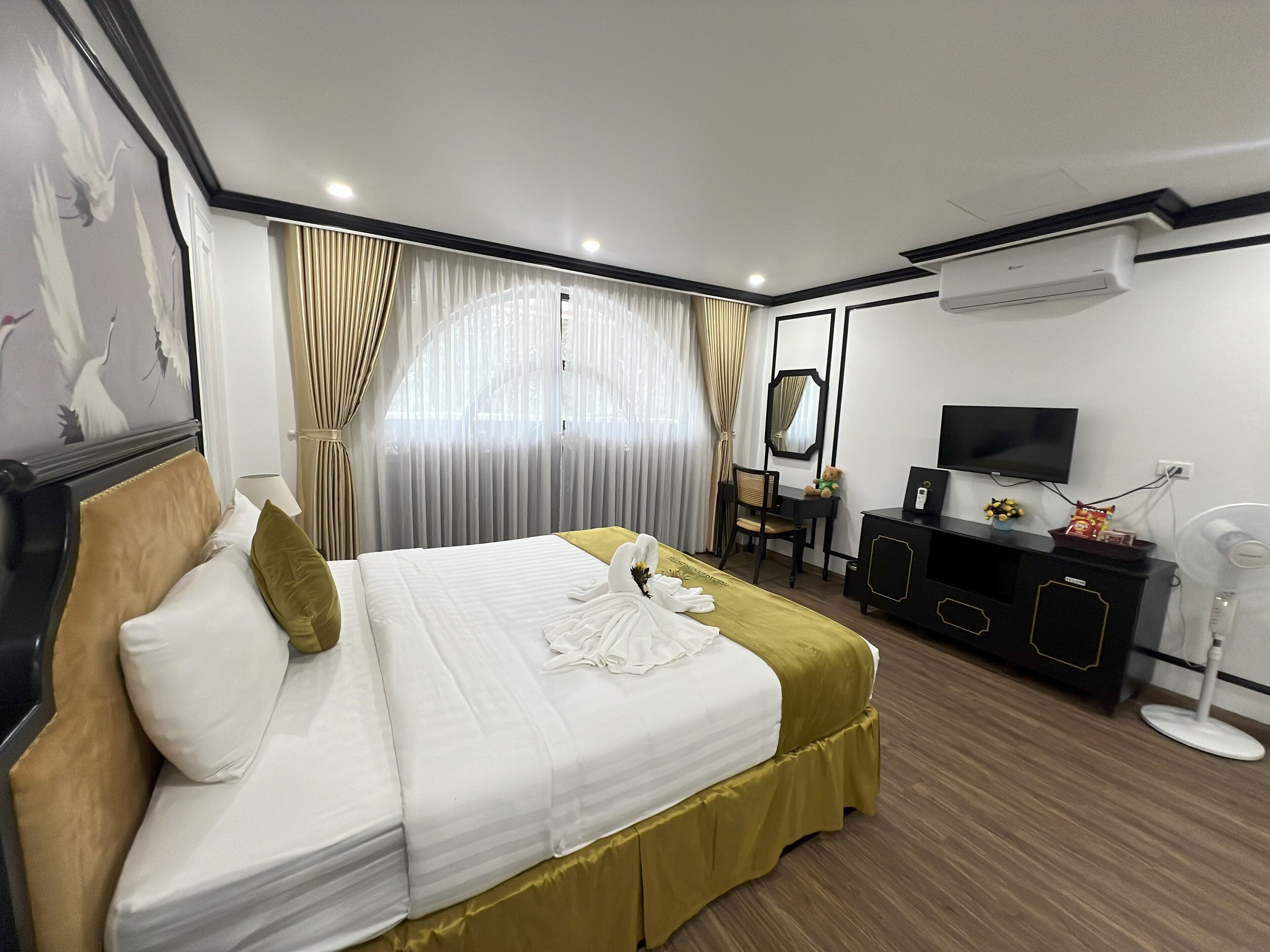A hotel room with a neatly made bed, a desk with a chair, a television, and an air conditioning unit.