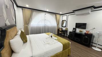 A hotel room with a neatly made bed, a desk with a chair, a television, and an air conditioning unit.