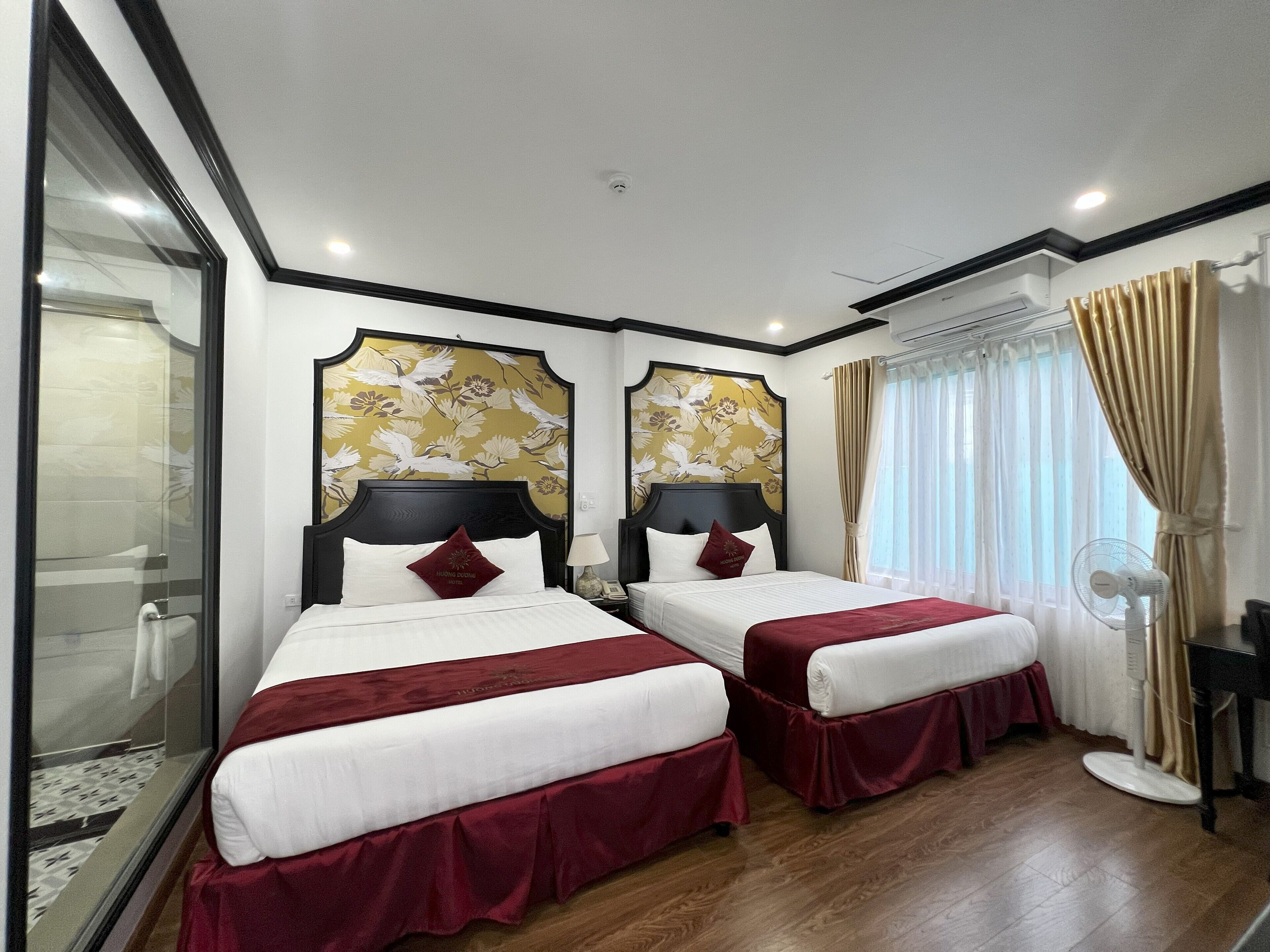 A hotel room with two beds, a large window with curtains, a standing fan, and a shower area.