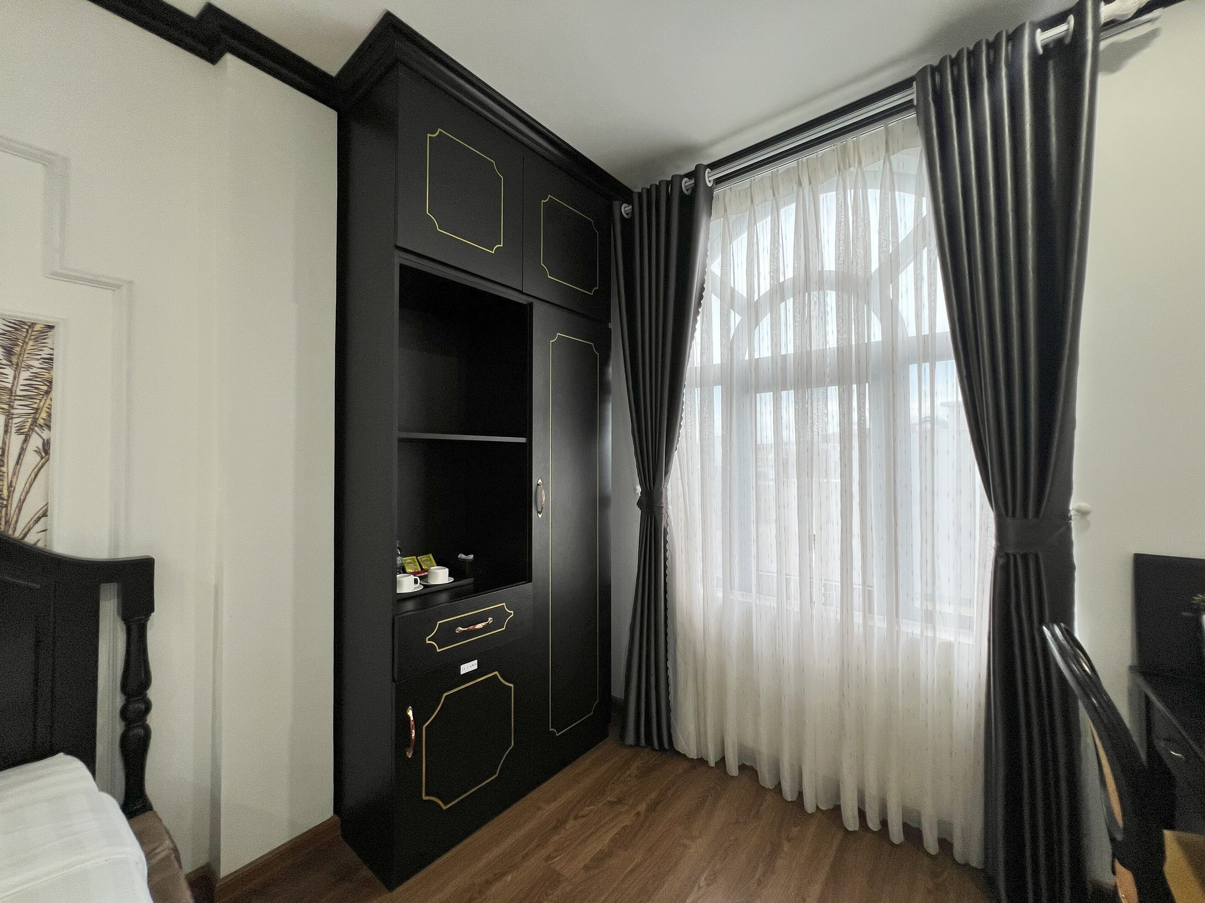 A room with a black wardrobe, a wooden floor, and a large window with sheer curtains.