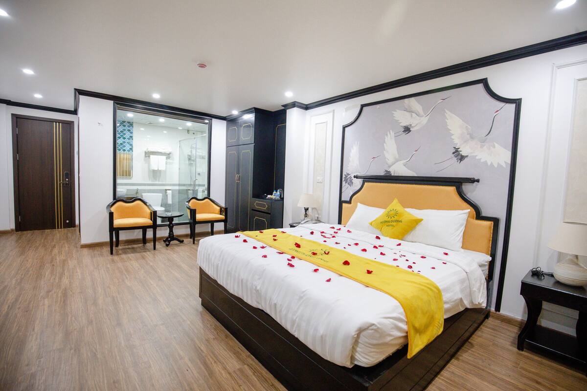 Quarto de hotel com uma cama grande, área de estar com mesa e cadeiras, um espelho e um quadro de um pássaro na parede.
