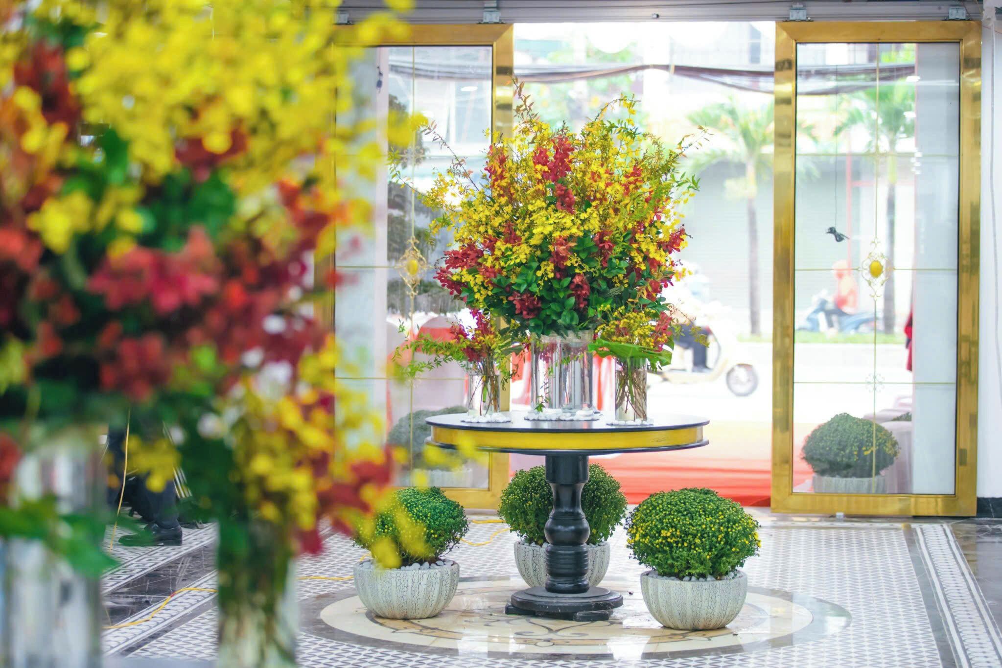 A lobby with a central round table surrounded by potted plants, a red carpet, and a glass door leading outside.