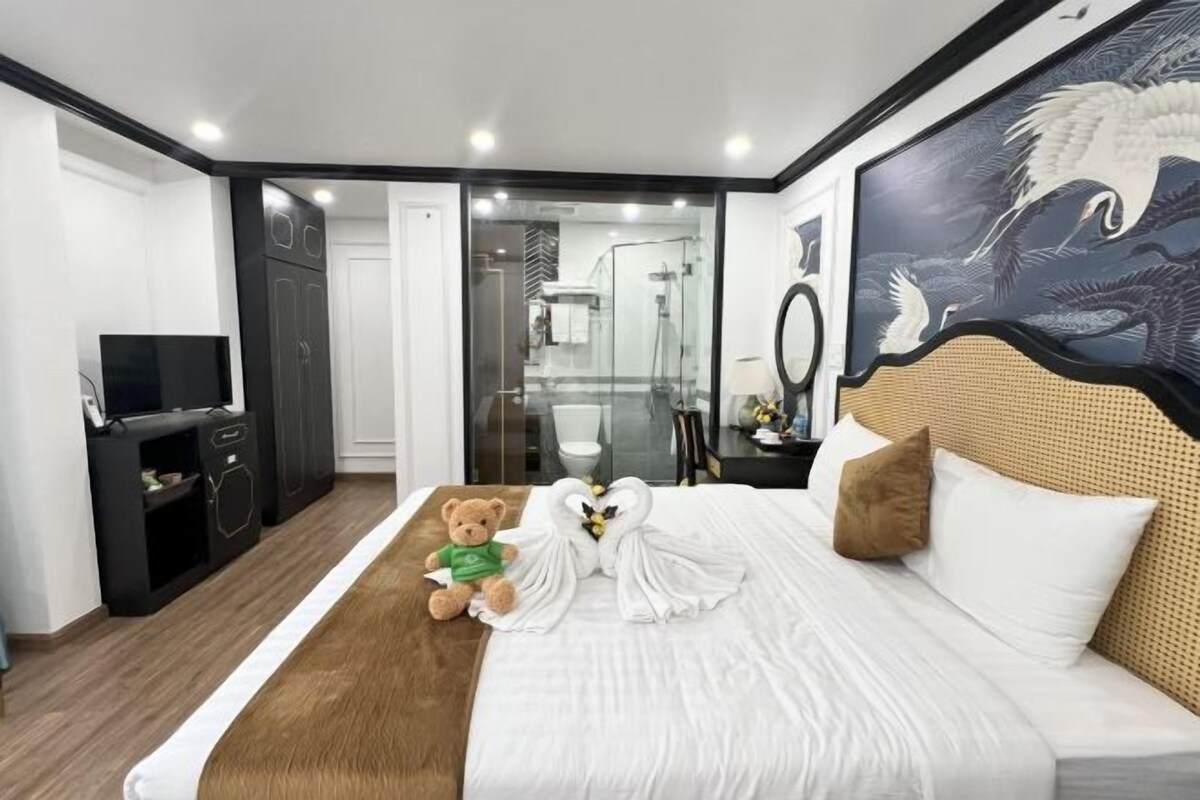 Une chambre d’hôtel avec un lit, un ours en peluche, une sculpture faite avec une serviette, une télévision et une salle de bain visible à travers une porte en verre.