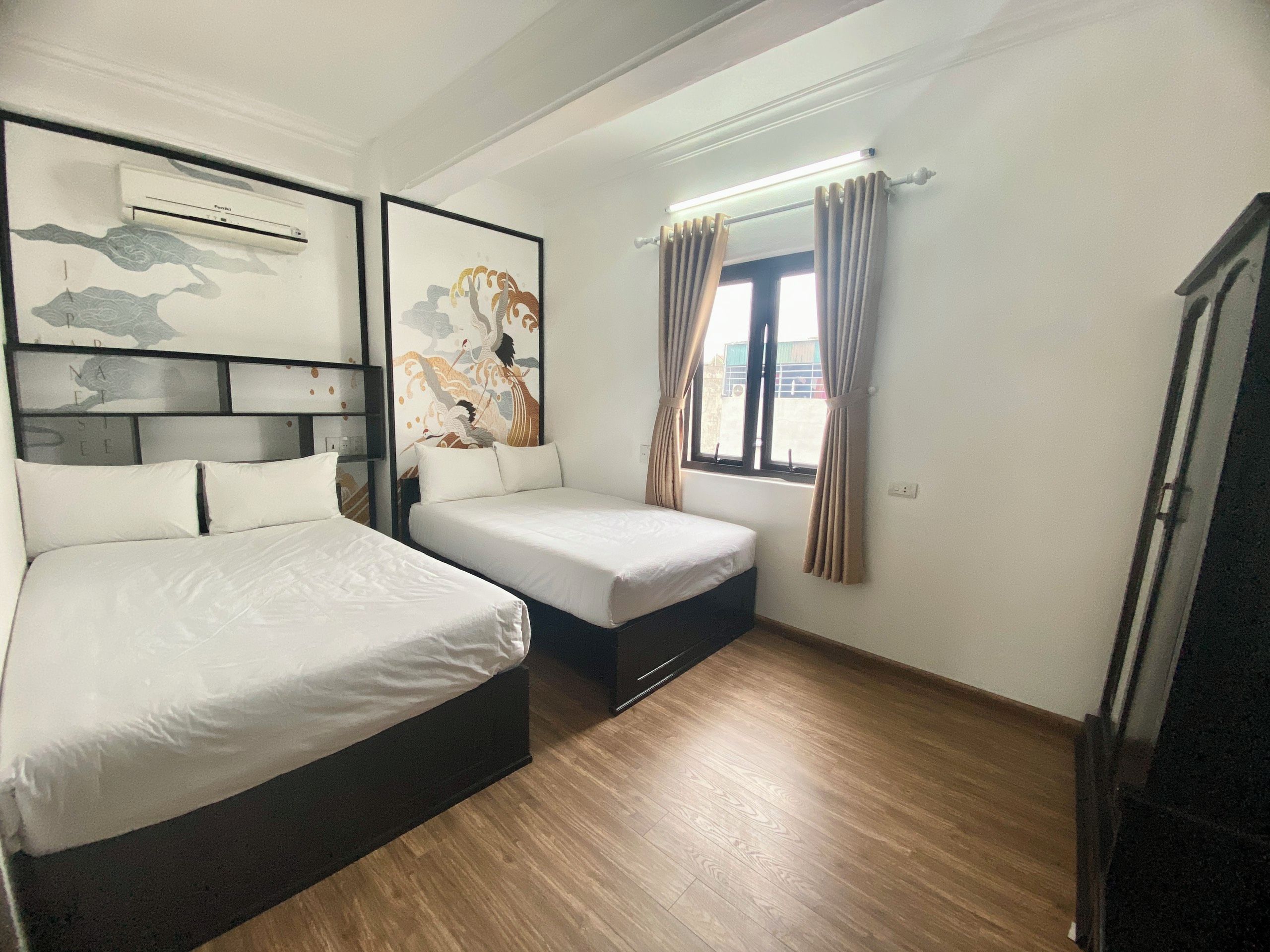 Ein Hotelzimmer mit zwei Betten, einer Wandklimaanlage, einem Fenster mit Vorhängen und einem Holzboden.