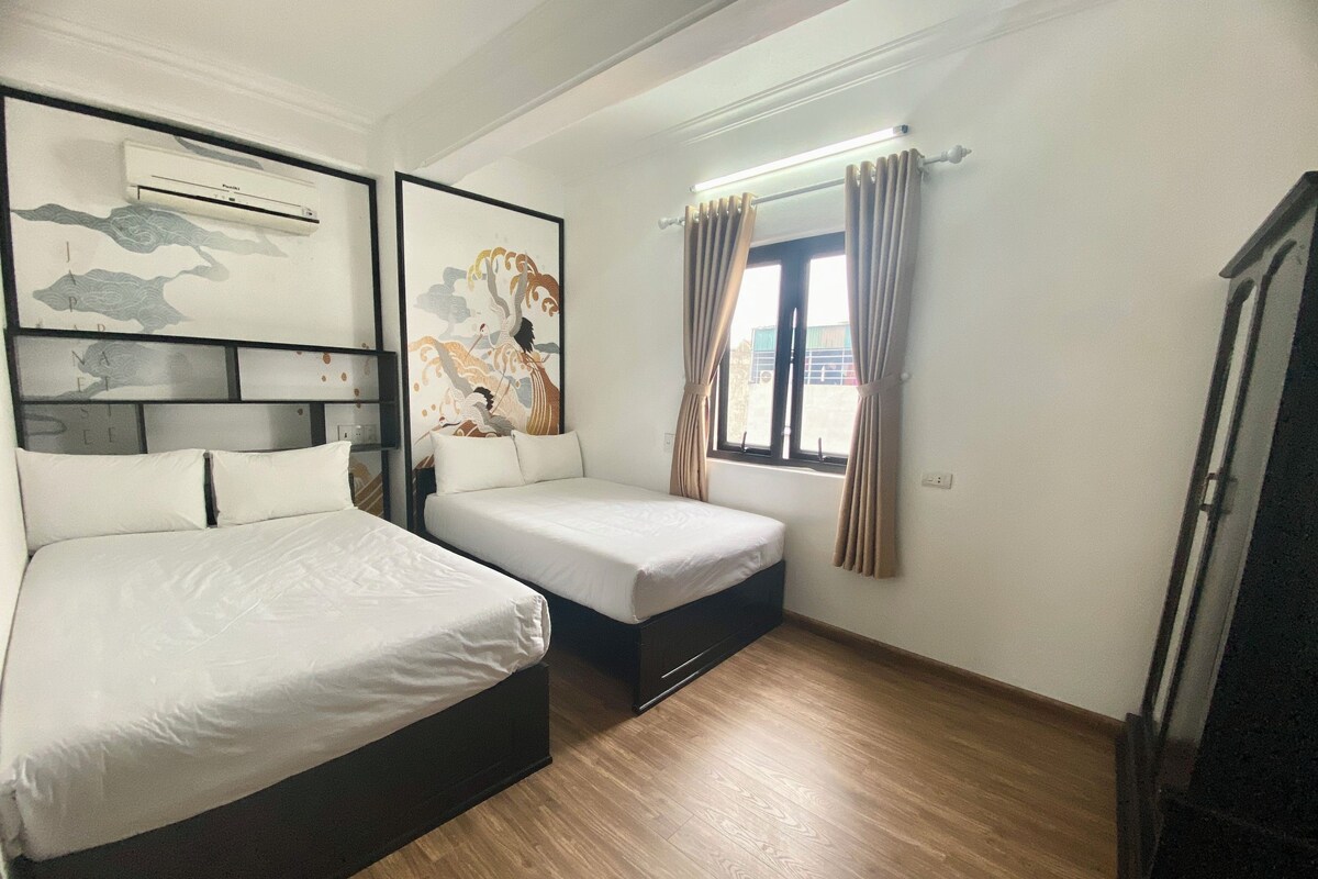 Ein Hotelzimmer mit zwei Betten, einer Wandklimaanlage, einem Fenster mit Vorhängen und einem Holzboden.