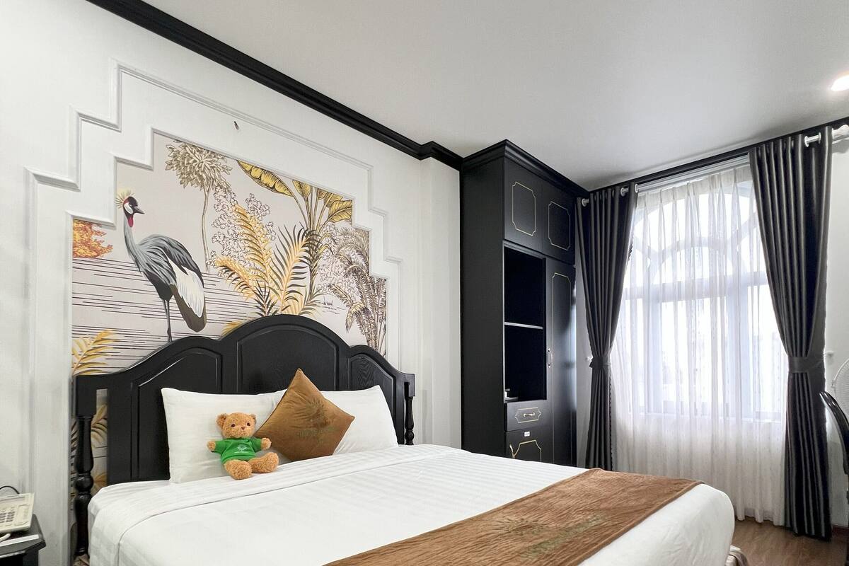 Cama bem arrumada com colcha marrom, travesseiro branco e um urso de pelúcia. Há uma parede com um quadro de tema natural acima da cama.