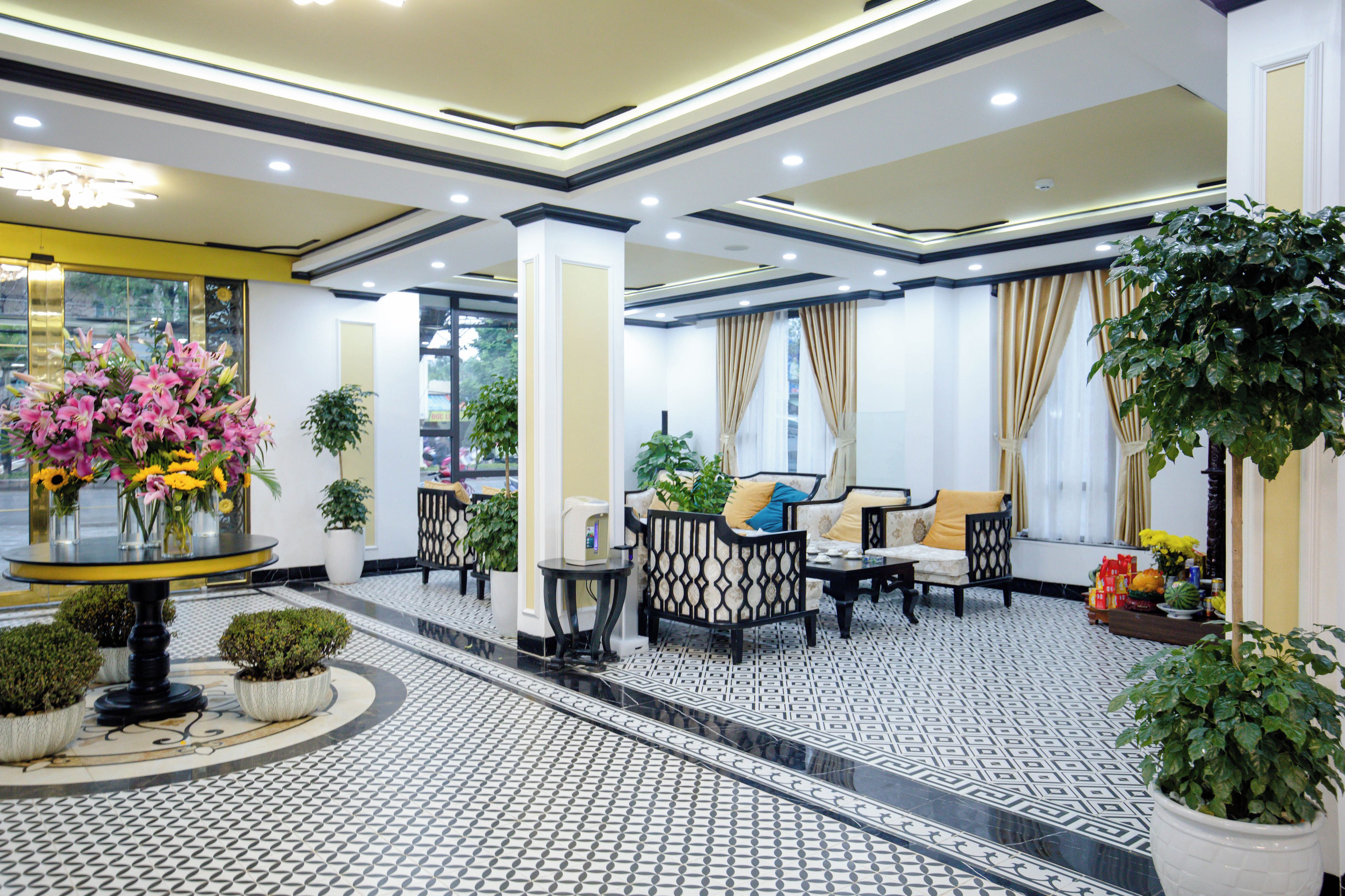 Un hall de hotel con suelo de baldosas, zona de estar con sofás y mesas, y plantas en macetas.