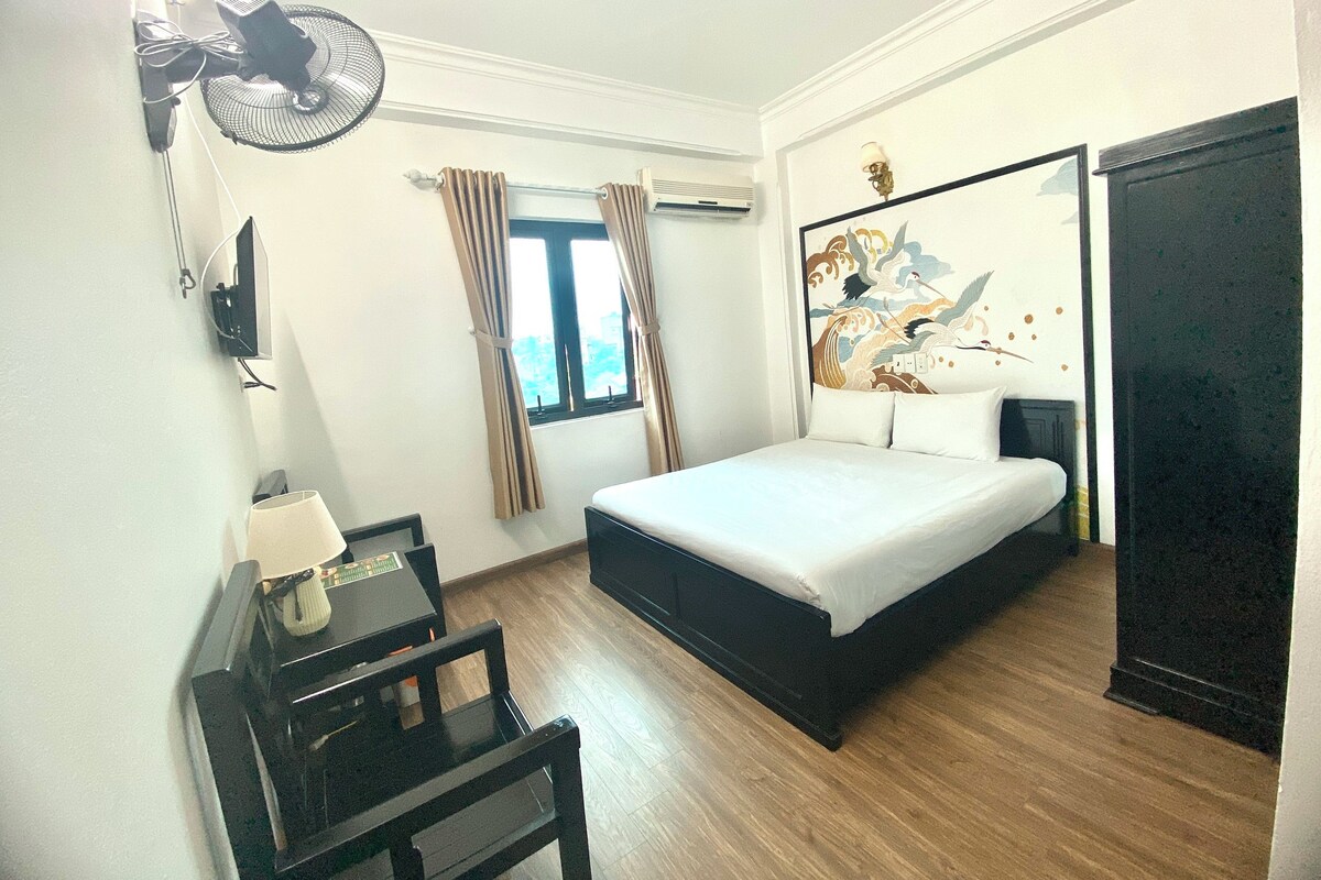 Ein Hotelzimmer mit einem Bett, einem Schreibtisch, einem Sessel, einem Ventilator, einem Fenster mit Vorhängen und einer Wand-Klimaanlage.