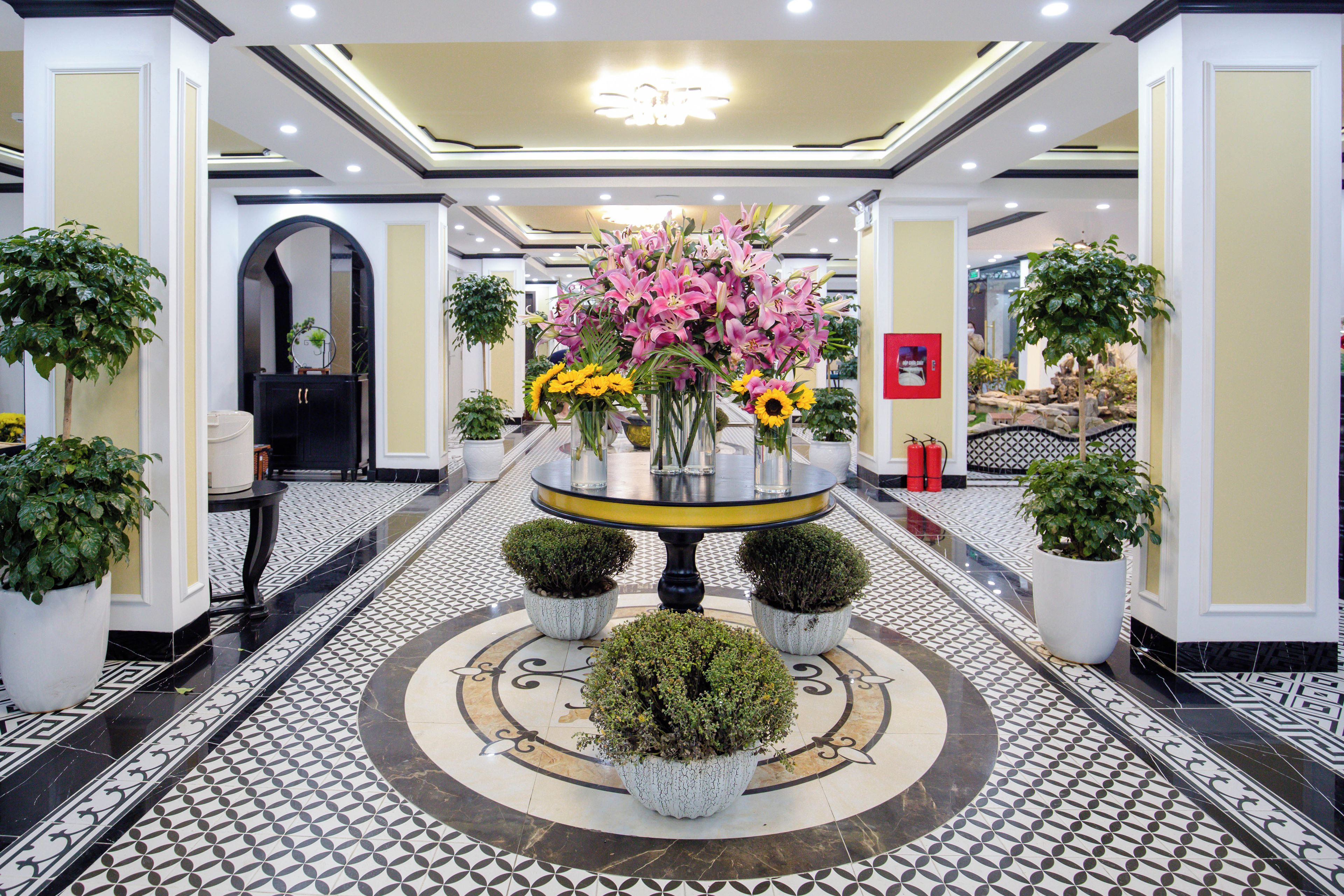 A hotel lobby with a central round table surrounded by potted plants and flowers.