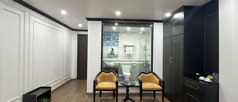 A modern hotel room with a wooden floor, a glass partition, two chairs, a small table, and a black wardrobe.