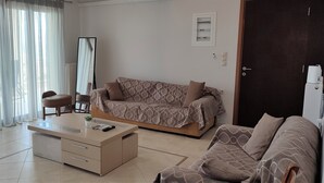 A modern living room with a sofa, a coffee table, and a chair. There is a large window with sheer curtains, a floor lamp, and a door with a refrigerator.