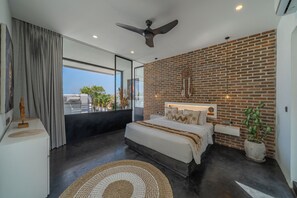 A modern bedroom with a large bed, a brick wall, a ceiling fan, and a view of the city. - Loft Erika (Canggu)