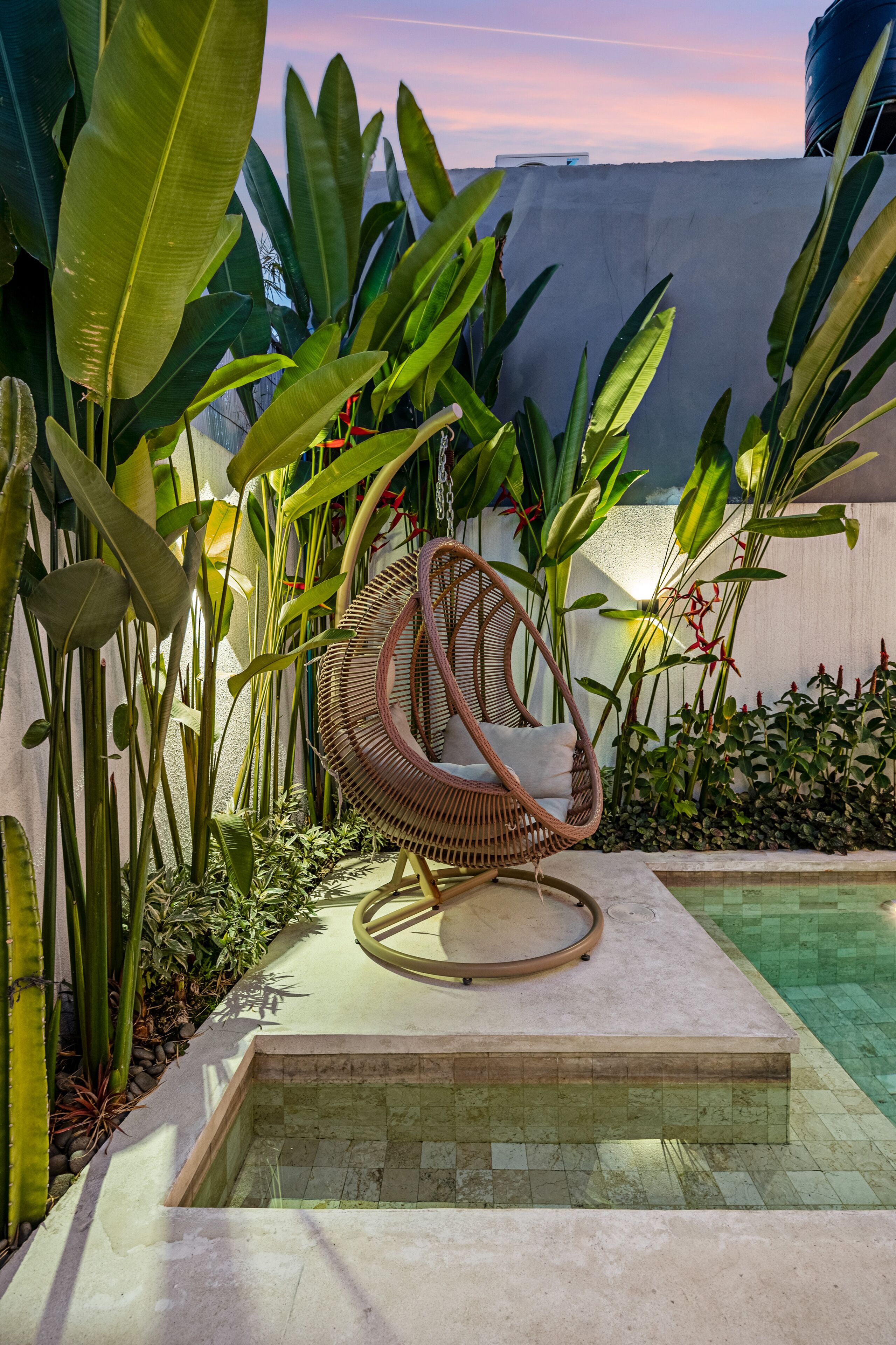 Ein Korbstuhl am Pool, umgeben von tropischen Pflanzen und einer Betonkante.
