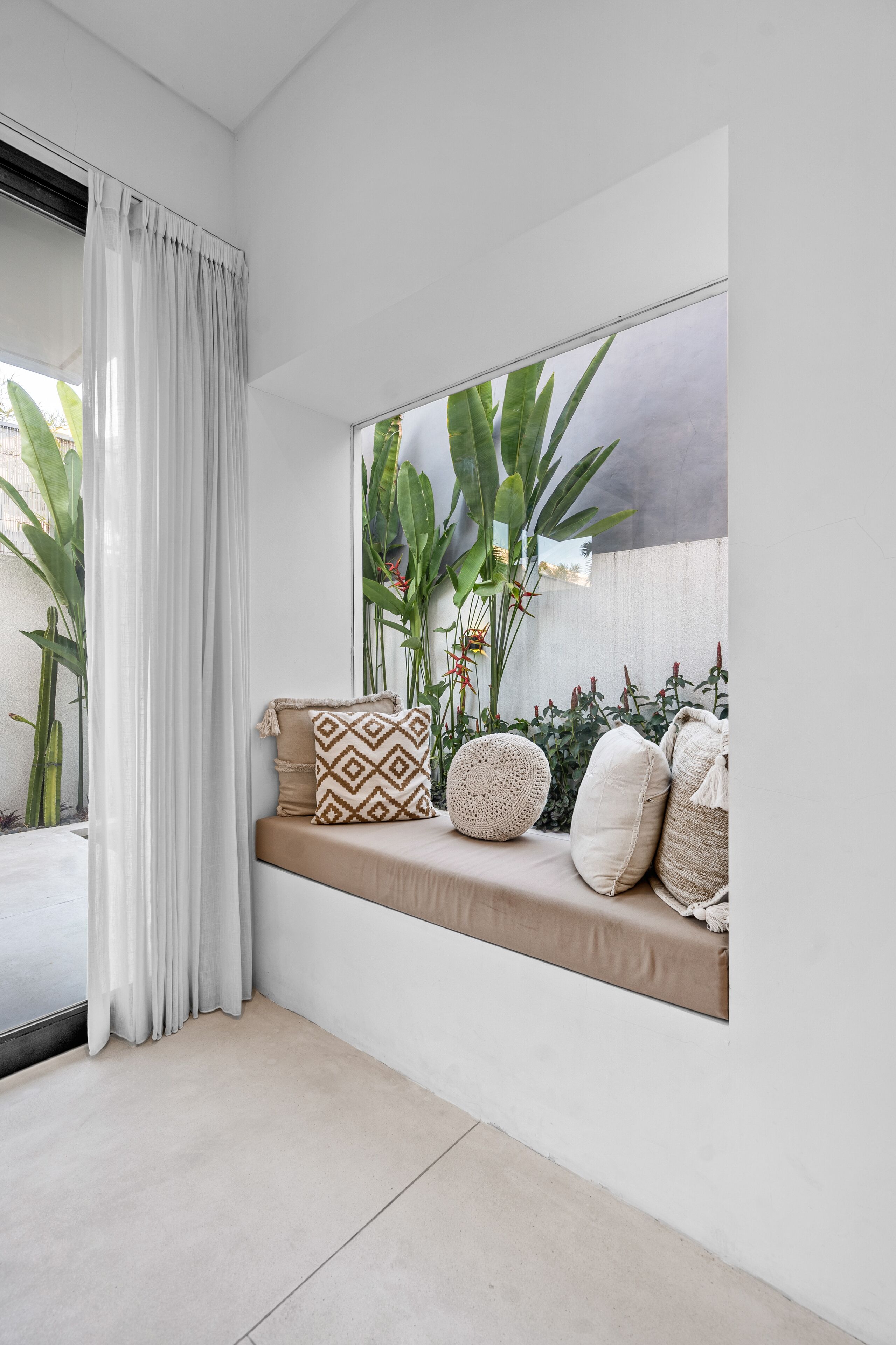 A room with a window seat, white curtains, and a view of greenery outside.