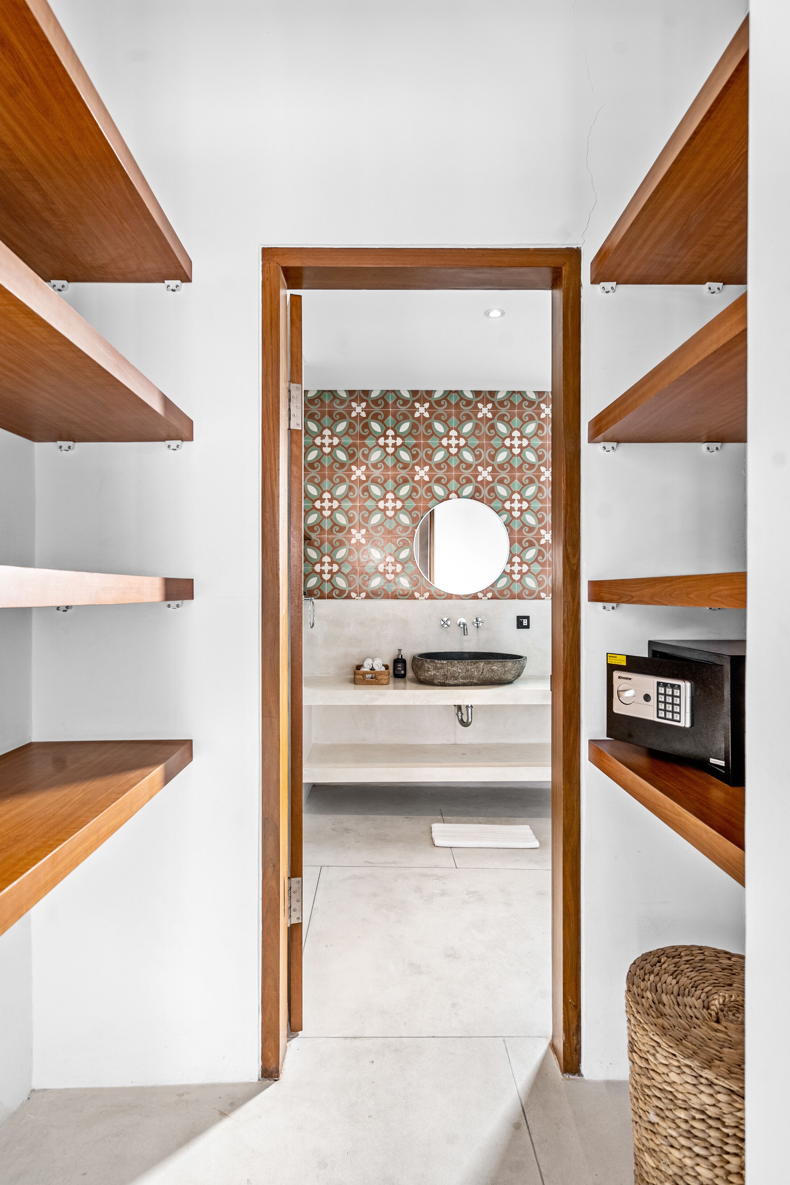 A narrow hallway with wooden shelves on either side, leading to a bathroom with a tiled backsplash, a round mirror, and a sink.