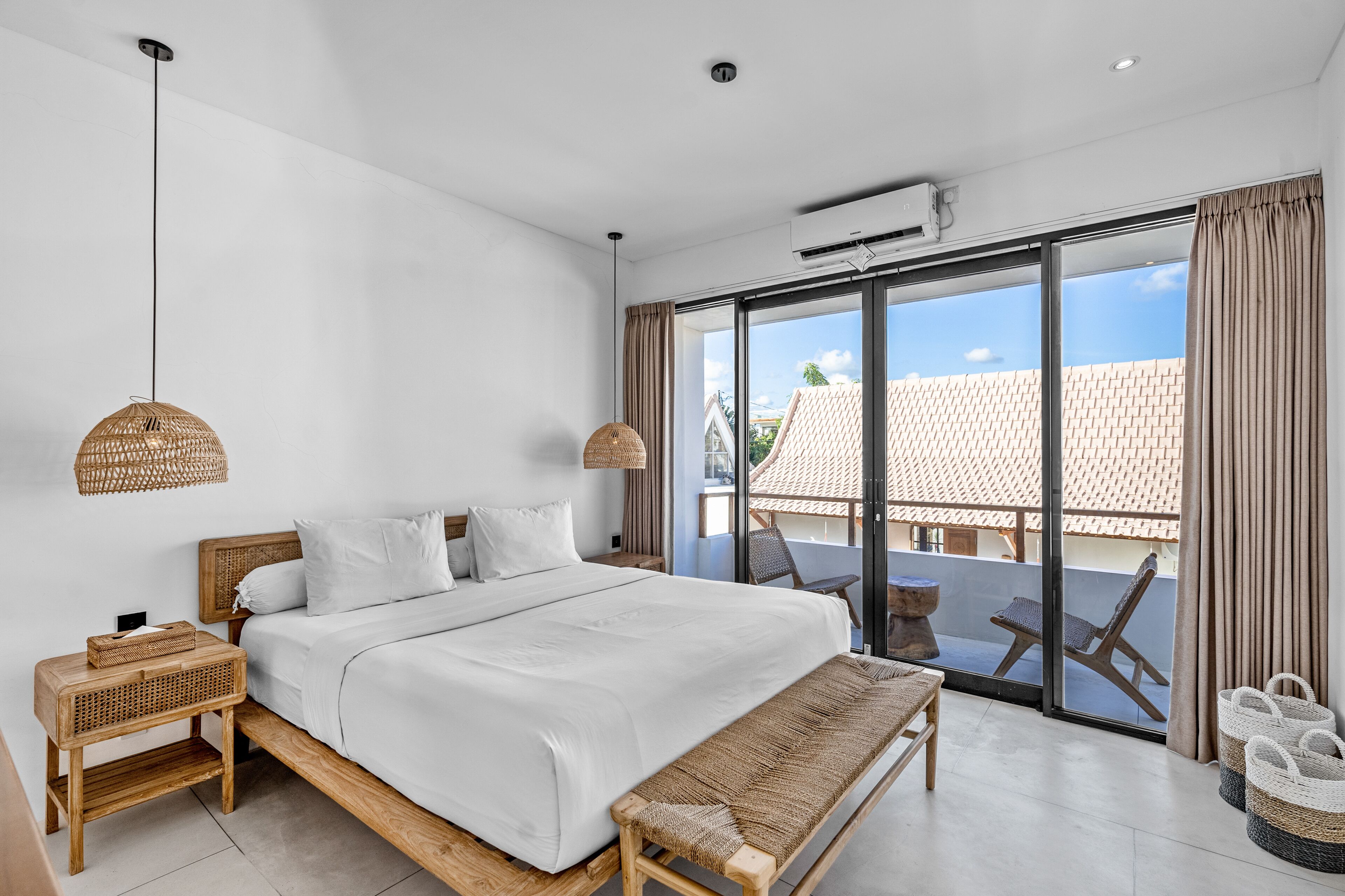A modern bedroom with a large bed, a wooden bench, wicker side tables, and a balcony with a view of a building.
