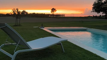 Un espace aménagé au bord de la piscine, avec un fauteuil de relaxation et une vue dégagée sur le coucher de soleil au-dessus d’un champ verdoyant.